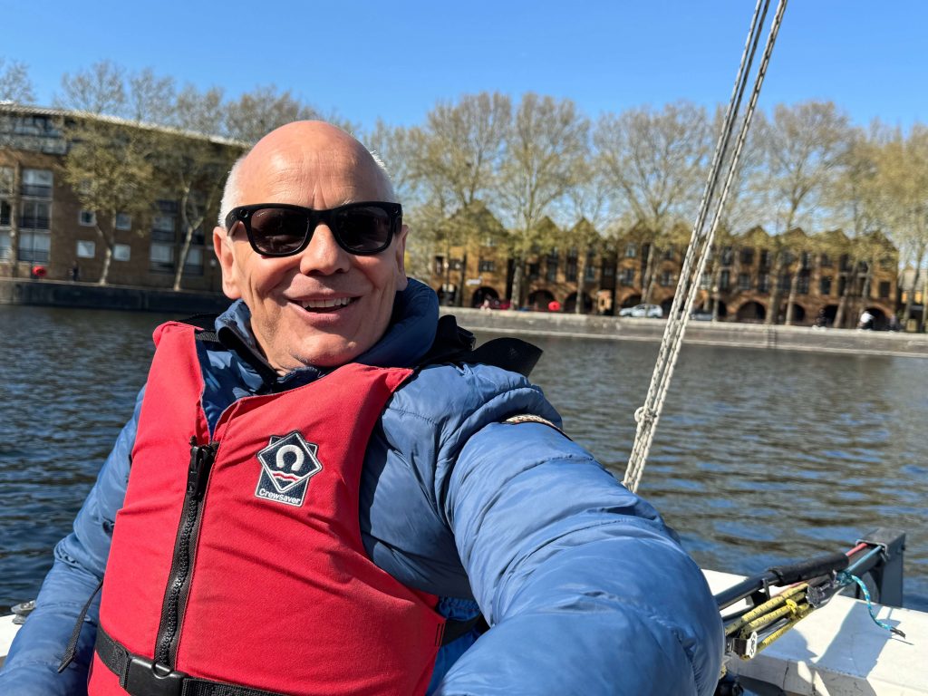 Man smiling on a boat wearing black sunglasses, blue jacket, and red Crewsaver life vest, with water, London-style brick buildings, and trees in the background on a sunny day.
