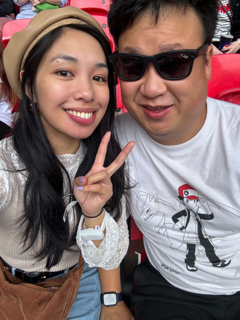 Woman and man smiling at the camera while sitting in red seats at Wembley Stadium in London. The woman is wearing a beige beret, lace top, and making a peace sign. The man is wearing black sunglasses and a white T-shirt with a cartoon character and dragon design.